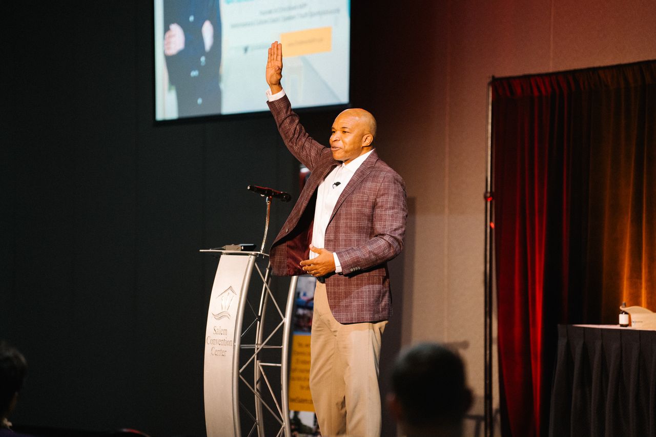 Chris Rivers raising his hand while speaking beside a lectern at the Salem Convention Center, engaging the audience mid-presentation.
