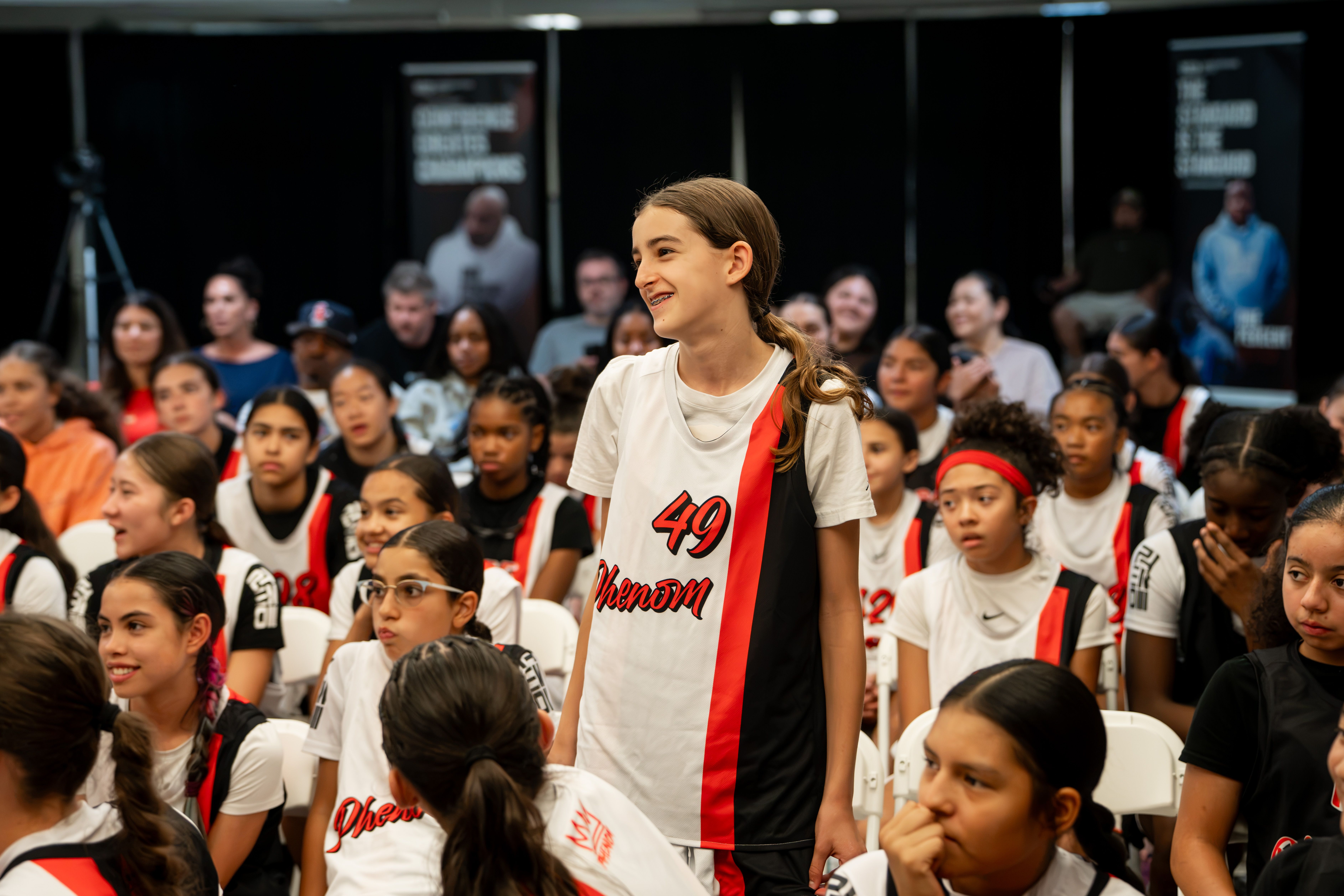 A young participant standing and smiling among a seated audience, the visible focal point of the room.